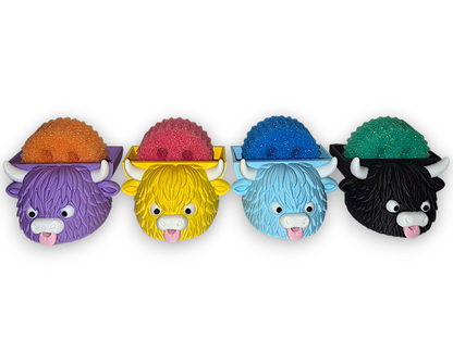 Four colorful highland cow-shaped soap dishes lined up on a white background, each holding a glittery sponge in a different color: purple with orange, yellow with pink, light blue with blue, and black with green.
