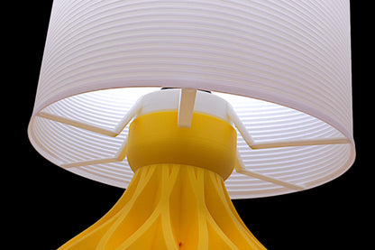 Table lamp – white striped cylindrical lampshade with sculptural yellow petal-like base, shown glowing

