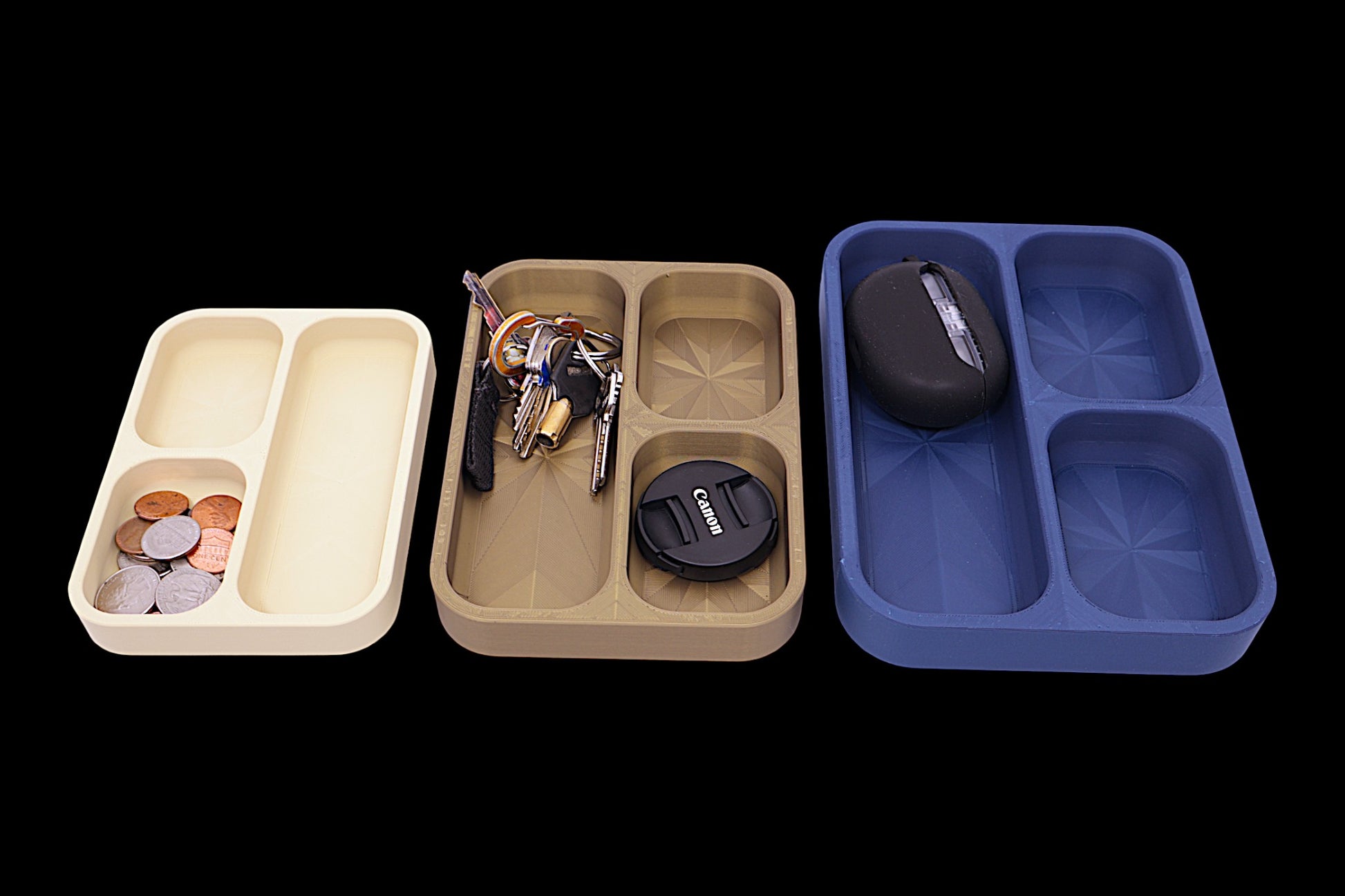 Three compartmentalized trays with various items including coins, keys, and a camera lens cap on a black background.