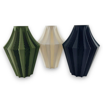 Three geometric vases are displayed side by side. The vases have a unique, angular design with multiple layers creating a ribbed effect. The vase on the left is olive green, the middle vase is off-white, and the vase on the right is Matte charcoal.